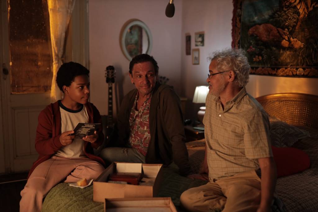 dois homens e uma criança conversando em cena da série com luz laranja todos em cima da cama