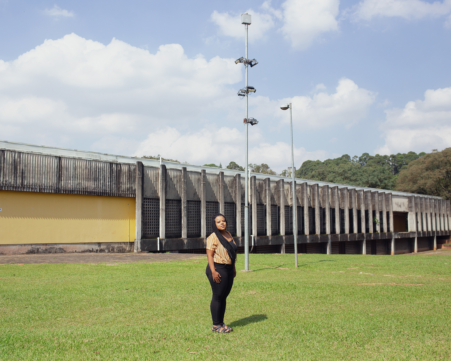 Luana Lopes em frente de um prédio amarelo e de cimento ao fundo, com grama e o poste
