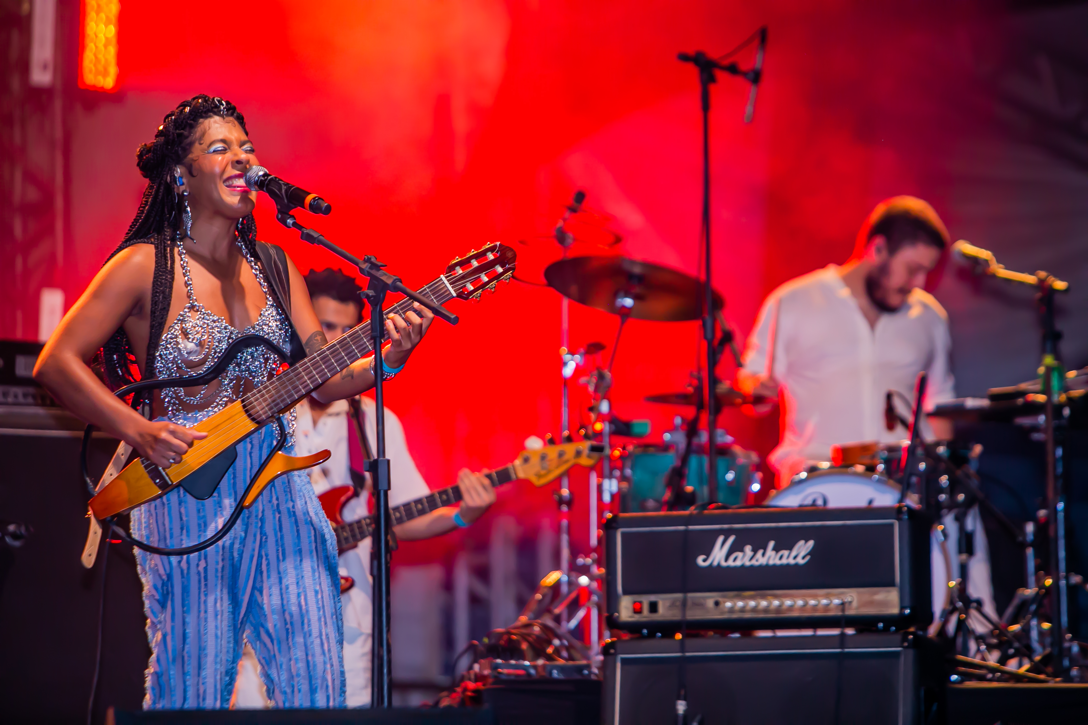 Josyara canta com violão na mão, atrás dela luz vermelha e dois músicos, guitarrista e baterista.